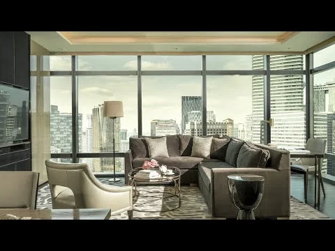Living features: L-shape sectional sofa in dark grey, glass coffee table, area rug with geometric pattern, floor-to-ceiling windows, city skyline view, wall-mounted TV, decorative wall paneling, recessed ceiling lights, light-colored walls, dark wood flooring; style: modern; view: city.
