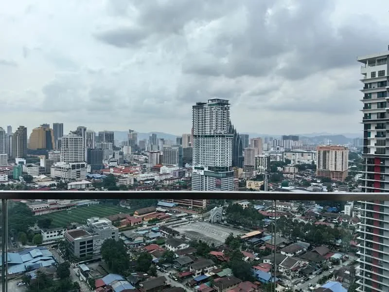 Exterior type: high-rise condominium; features: city skyline view, glass balcony railing, surrounding urban landscape, mixed-use buildings, residential and commercial structures, green spaces visible, cloudy sky; style: modern.