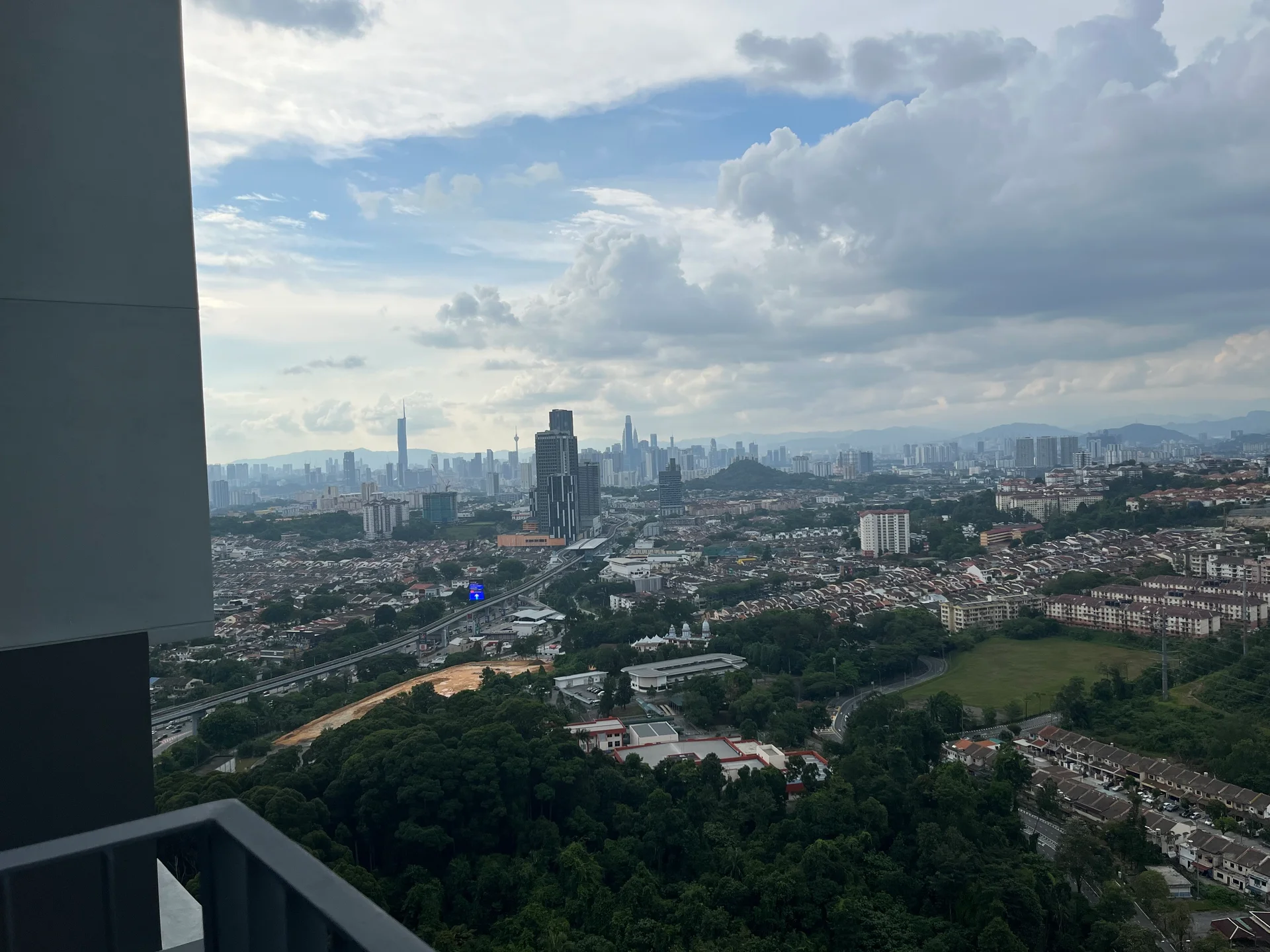 Exterior type: high-rise condominium; features: city skyline view, surrounded by greenery, urban landscape, high-rise buildings in the background, road visible in the foreground, hills in the distance; style: modern.