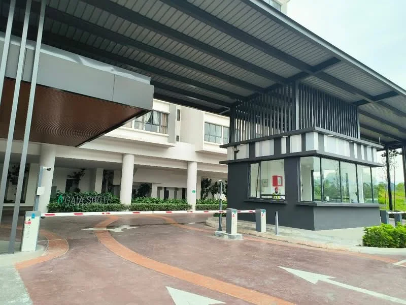 Exterior type: main entrance gate; features: covered entrance with metal roofing, security guard house, automatic boom gate, paved driveway, landscaped greenery, modern architectural design, glass windows on guard house, signage visible on facade; style: modern.