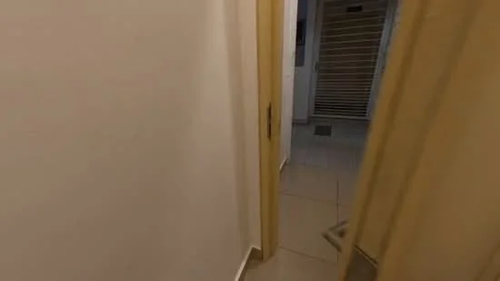 Room features: white painted walls, tile flooring, wooden door, door frame, light switch, visible corridor, sliding door at end of corridor.