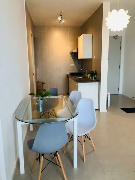 Dining features: glass dining table, 4 dining chairs in grey and white, pendant light fixture, light beige tiled flooring, white walls, kitchenette visible in background, wall-mounted cabinets, dark countertop, small indoor plant on table, recessed ceiling lights; style: modern. Kitchen features: white cabinetry, dark countertop, stainless-steel sink, built-in stove, wall-mounted cabinets; style: modern.