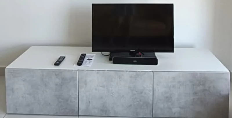 Living features: white TV console with grey drawers, flat-screen TV, remote controls, minimalist design, light-colored flooring, neutral wall color; style: modern.