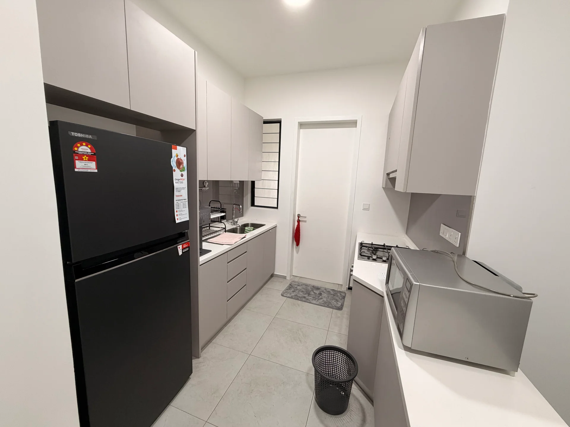 Kitchen features: grey cabinetry, black refrigerator, built-in gas stove, microwave, stainless-steel sink, white countertop, tile flooring, white walls, window with grid design, kitchen towel hanging on door, waste basket; style: modern.