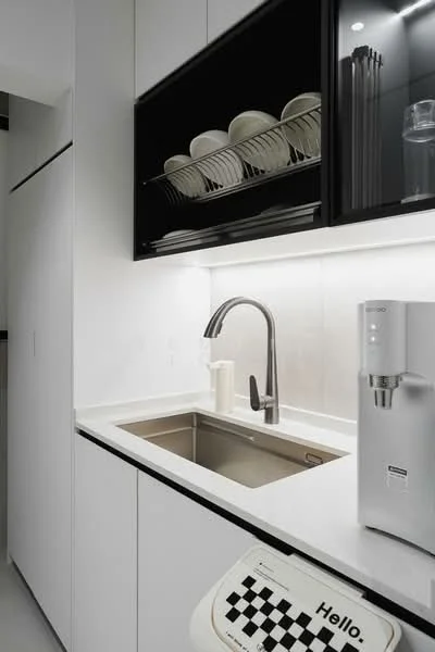 Kitchen features: single-bowl stainless-steel sink, chrome pull-down faucet, white quartz countertop, black cabinetry, glass-front upper cabinets, white plates on display, built-in water dispenser, integrated refrigerator, minimalist design, LED under-cabinet lighting; style: modern.