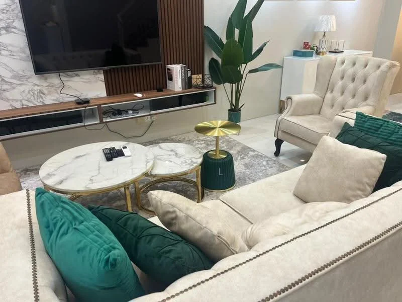 Living features: beige fabric sofa with decorative cushions, 2-seater beige armchair, marble coffee table with gold accents, green accent table, flat-screen TV mounted on wall, wooden TV console with storage, decorative plant in corner, area rug with geometric pattern, recessed lighting, wall paneling with wood finish, light-colored tiled flooring; style: modern.