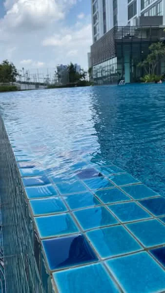 Exterior type: swimming pool deck; features: infinity pool, blue mosaic tile, water surface reflecting sky, surrounded by greenery, deck area with loungers, modern architecture in background; style: modern.
