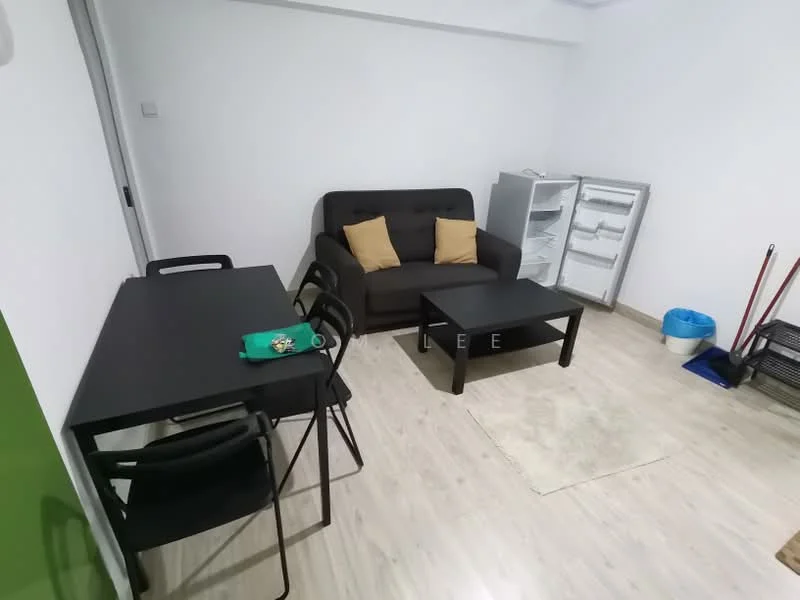 Living features: grey fabric sofa, 2 beige throw pillows, black rectangular coffee table, black dining table for 4, 4 black dining chairs, light wood laminate flooring, white painted walls, refrigerator, blue plastic trash bin, cleaning tools in corner; style: modern.