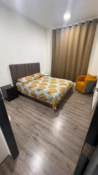Bedroom features: queen-size bed with floral bedding, upholstered headboard, wooden bedside table, laminate flooring, curtains in brown, armchair in orange and grey, light fixture on ceiling, white painted walls; style: modern.