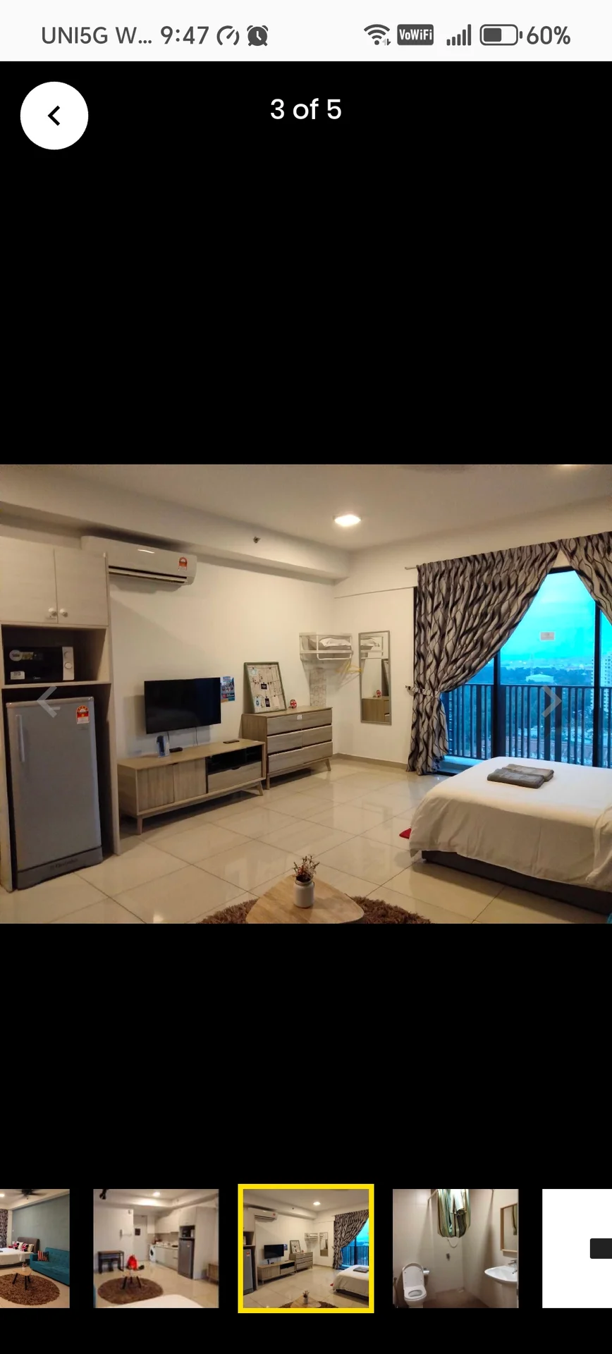Bedroom features: queen-size bed with white bedding, 2 bedside lamps, wall-mounted TV, wooden dresser with drawers, fridge, floor-to-ceiling windows, curtains with floral pattern, air conditioning unit, tile flooring, decorative area rug; style: modern; view: city.