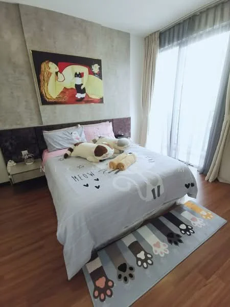 Bedroom features: queen-size bed with grey bedding, upholstered headboard, decorative wall art with a woman and cats, wooden flooring, floor-to-ceiling windows with sheer curtains, light-colored curtains, bedside table, area rug with paw print design, stuffed animals on the bed; style: modern.