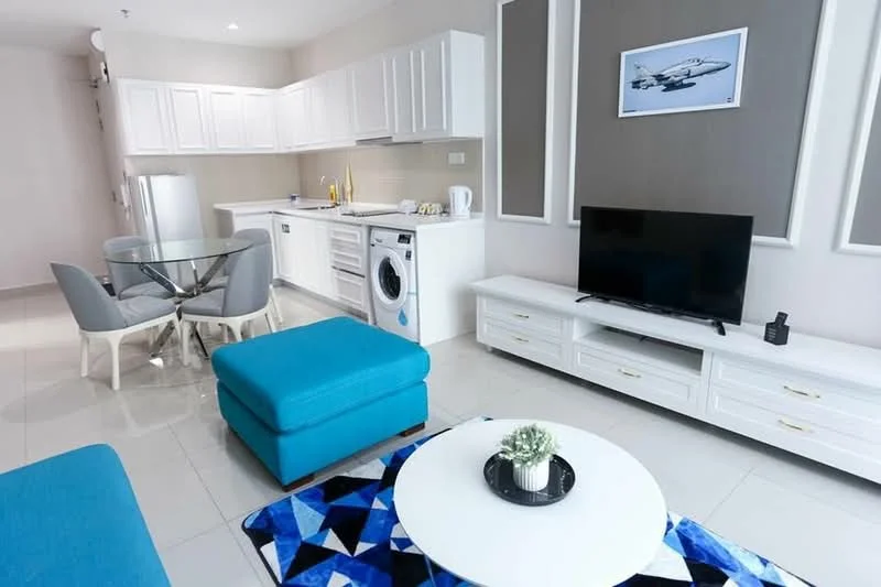 Living features: white cabinetry, grey accent wall, sofa in blue, coffee table with circular top, TV console in white, wall-mounted TV, area rug with geometric pattern, dining table with glass top, 4 dining chairs, washing machine, refrigerator, recessed downlights, tile flooring, plant in ceramic pot; style: modern.