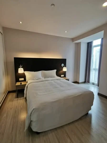 Bedroom features: queen-size bed, upholstered headboard, two bedside lamps, wooden bedside tables, light wood flooring, white bedding, neutral wall colour, floor-to-ceiling window, curtains, recessed lighting; style: modern; view: city.
