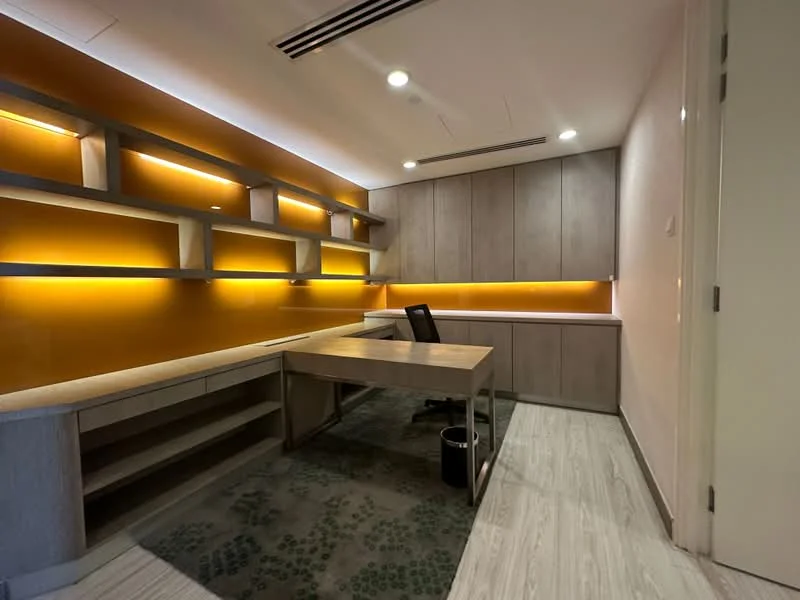 Study features: wooden desk, black office chair, built-in cabinetry, shelving with backlighting, light wood cabinetry, green patterned carpet, recessed ceiling lights, orange accent wall, white painted walls; style: modern.