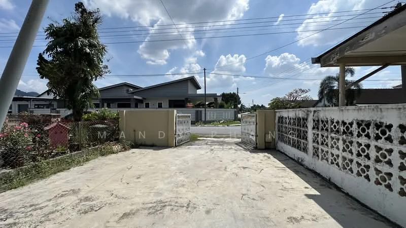 Exterior features: open gate entrance, concrete driveway, fenced perimeter, landscaped garden with shrubs, mature trees, single-storey building in background, clear blue sky, power lines visible; style: modern.