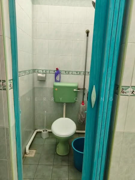 Bathroom features: green toilet, white toilet bowl, green toilet tank, white tiled walls, green accent wall, grey floor tiles, shower head, mop, cleaning supplies, blue bucket, toilet paper holder, floor drain.