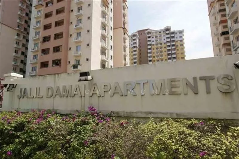 Exterior type: low-rise apartment block; features: signage with 'Jalil Damai Apartments', landscaped garden with flowering plants, multiple residential buildings, balconies visible on buildings, cream and pastel-colored facade, surrounding greenery; style: modern.