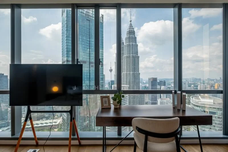 Study features: large floor-to-ceiling windows, wooden desk, white upholstered chair, TV on stand, decorative plant, city skyline view, natural light, wooden flooring; style: modern; view: city.