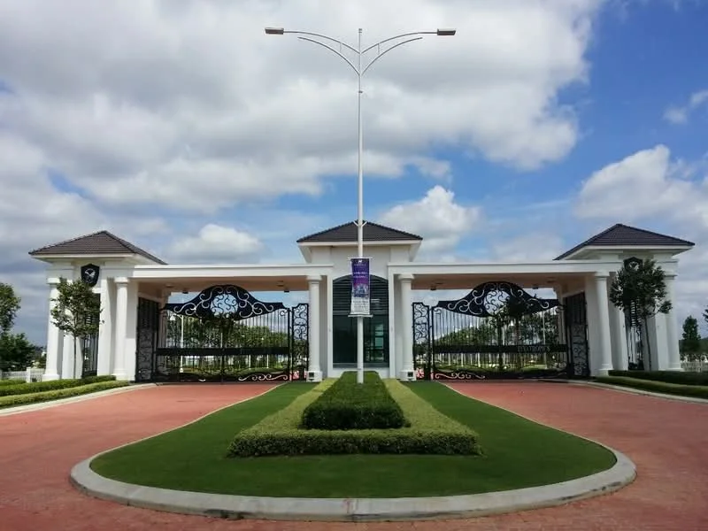 Exterior type: main entrance gate; features: ornate wrought iron gates, white painted facade, two symmetrical gatehouses, flat roof with overhang, manicured lawn, landscaped garden beds, paved driveway, street lamp, clear blue sky, clouds; style: modern.
