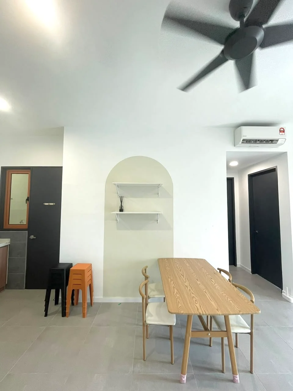 Dining features: wood dining table, 4 dining chairs, open shelving on wall, grey tiled flooring, white painted walls, black wall-mounted split AC, 5-blade ceiling fan, dark door, light door, stool seating, arched wall feature; style: modern.