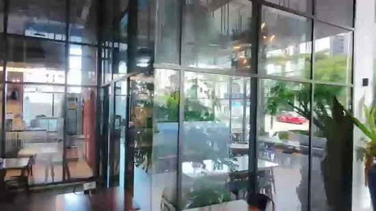 Room features: large glass windows, metal frame structure, wooden flooring, indoor plants, open layout, modern lighting fixtures, dining tables, chairs, decorative wall, visible outdoor view; style: modern; view: city.