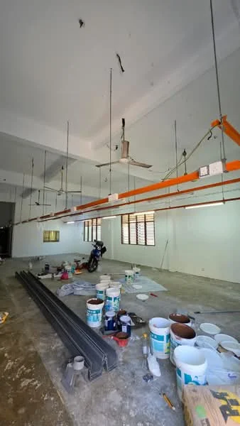 Room features: unfinished concrete flooring, white painted walls, exposed ceiling with hanging light fixtures, ceiling fan, multiple paint buckets, construction materials on the floor, window with wooden frame, open space layout.
