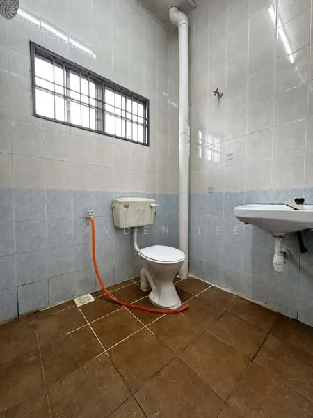 Bathroom features: white tiled walls, light blue tiled walls, brown tiled flooring, one-piece toilet, wall-mounted sink, exposed plumbing, window with metal grille, orange water supply line.