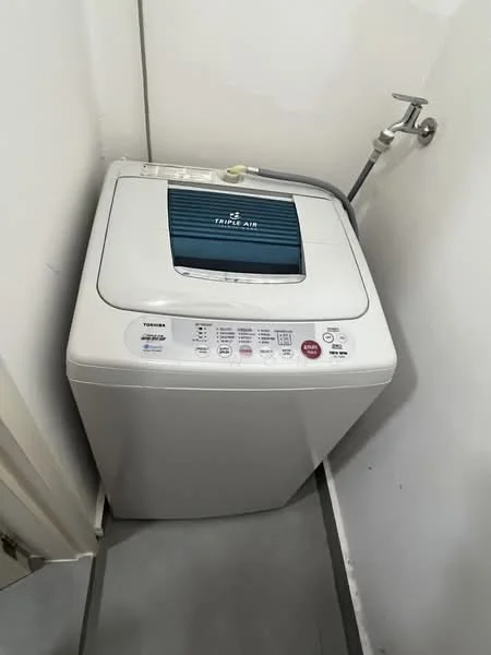Laundry features: top-load washing machine, white appliance, control panel with buttons, water inlet hose, drain hose, grey tiled flooring, corner placement, wall-mounted faucet.