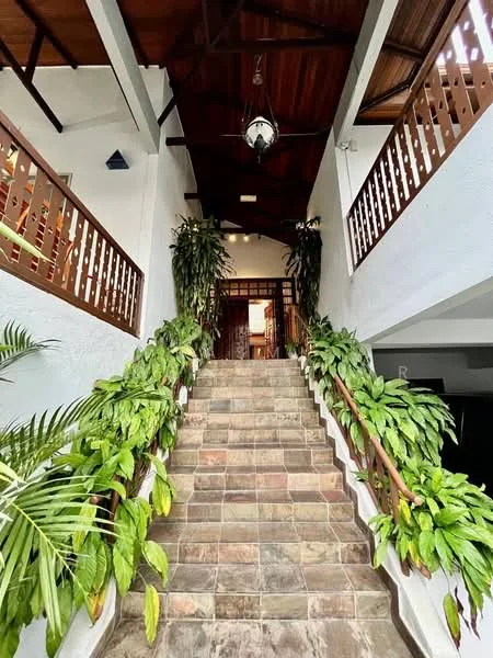 Room features: stone tile flooring, wooden staircase, high ceiling with exposed beams, ceiling fan, indoor plants, white painted walls, wooden railing, double wooden doors; style: tropical-modern; view: garden.