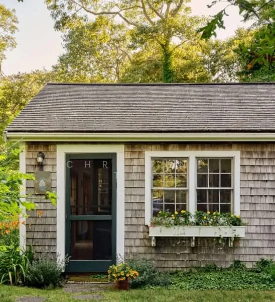 Exterior type: detached bungalow; features: shingle facade, green front door, two windows with white trim, window box with flowers, sloped roof, manicured garden, stone pathway, surrounded by trees; style: traditional.