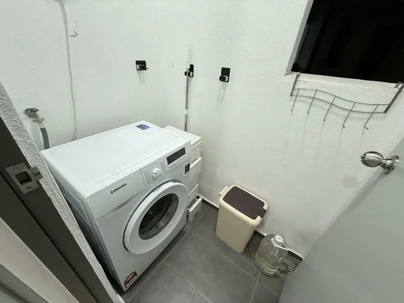 Laundry features: front-load washing machine, grey tile flooring, white painted walls, plastic laundry basket, wall-mounted storage hooks, small waste bin, door with handle.