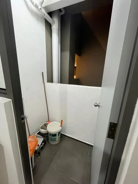 Room features: white painted walls, grey tiled flooring, pipework visible on wall, bucket, cleaning supplies, broom, small storage space, door leading to another area.