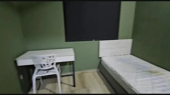 Bedroom features: green painted walls, wooden flooring, single bed, white bed frame, desk with white surface, metal legs on desk, plastic chair, black window blind.