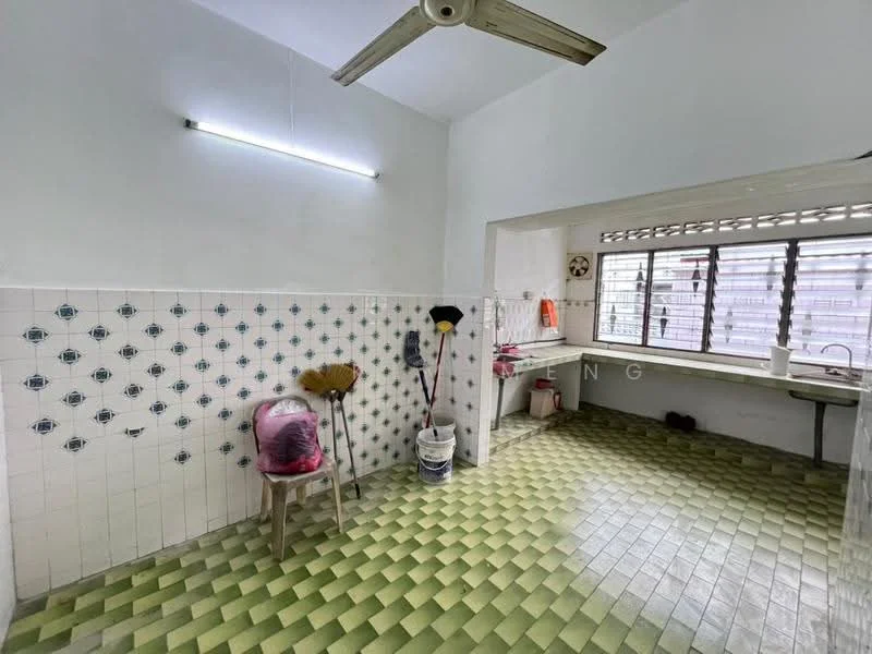 Kitchen features: green and yellow checkered tile flooring, white painted walls, exposed ceiling with fluorescent light, window with metal grille, sink with faucet, cleaning supplies including broom and mop, plastic chair, bucket, wall-mounted shelf.