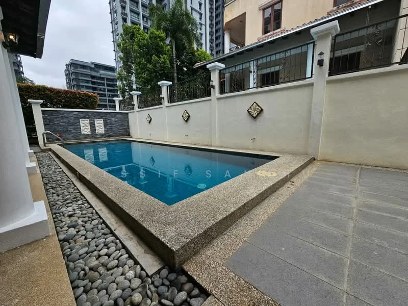 Exterior type: detached bungalow; features: infinity pool, pool deck with stone paving, landscaped garden with pebbled area, surrounded by low wall with decorative elements, mature trees in background, adjacent to multi-storey buildings; style: modern.