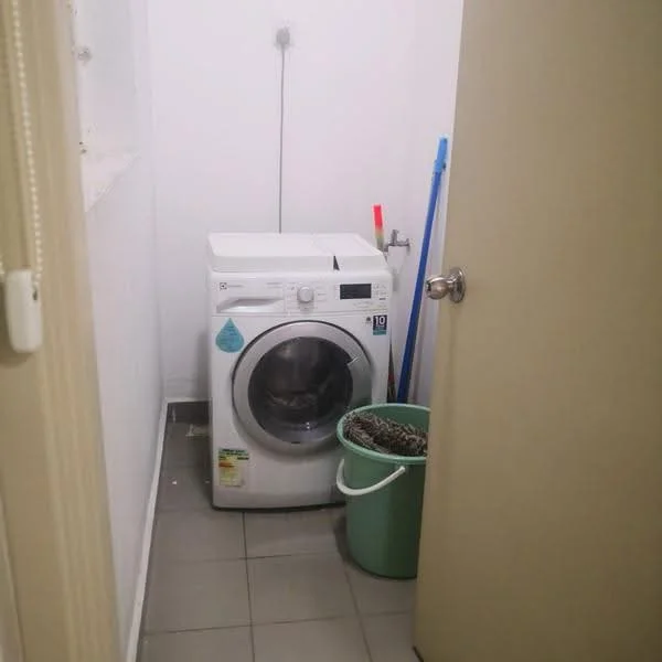 Laundry features: front-load washing machine, grey tiled flooring, white painted walls, green plastic laundry basket, cleaning supplies (broom and mop), wall-mounted water supply, light fixture on ceiling.