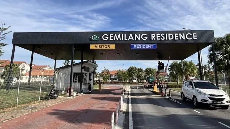 Exterior type: main entrance gate; features: guard house at entrance, visitor and resident lanes, security booth, paved driveway, landscaped greenery, signage for Gemilang Residence, surrounded by palm trees, fenced perimeter; style: modern.