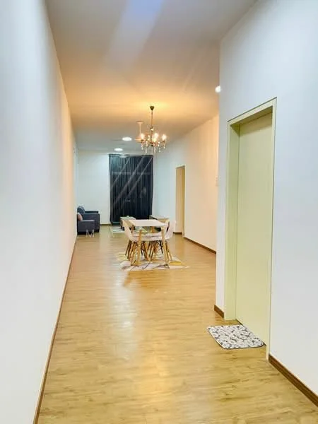 Room features: light wood laminate flooring, white painted walls, chandelier light fixture, dining table with white chairs, sofa in grey fabric, doorway leading to another room, doorway leading to elevator, decorative rug near doorway; style: modern.