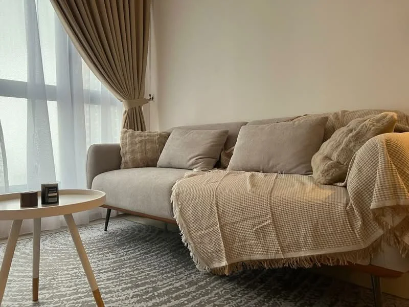 Living features: 3-seater fabric sofa in beige, decorative throw blanket, 2 decorative cushions, round coffee table with wooden legs, area rug with geometric pattern, floor-to-ceiling windows with sheer curtains, curtains in light brown, neutral painted walls; style: modern.