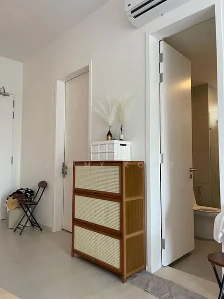 Room features: white painted walls, light grey flooring, wooden dresser with rattan detailing, white door, open door leading to bathroom, wall-mounted air conditioning unit, folding chair, decorative vase with dried plants; style: modern.