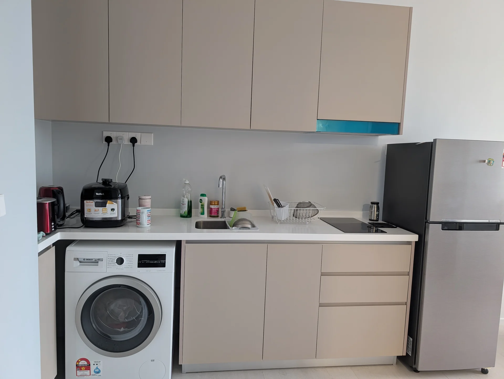 Kitchen features: light beige cabinetry, white countertop, single-bowl stainless-steel sink, chrome pull-down faucet, built-in refrigerator, front-load washing machine, black rice cooker, toaster, dish rack, cleaning supplies on countertop, wall-mounted electrical outlets; style: modern.
