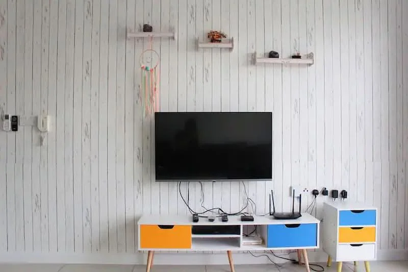 Living features: white wooden panelled walls, flat-screen TV, white TV console with blue and orange drawers, decorative shelves above TV, cable management visible, router on console, multiple power outlets, decorative items on shelves; style: modern.