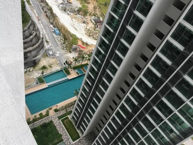 Exterior type: high-rise condominium; features: glass curtain wall facade, infinity pool, landscaped pool deck, surrounding greenery, construction site in background, paved walkway, outdoor seating area; style: modern.