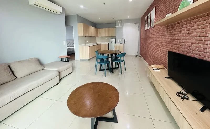 Living features: 3-seater fabric sofa in light grey, round wooden coffee table, TV console with wall-mounted TV, wooden shelving unit, white walls, grey accent wall with brick pattern, dining table with 4 blue chairs, ceramic tile flooring, air conditioning unit, recessed ceiling lights; style: modern. Kitchen features: kitchen cabinetry in light wood, white countertop, stainless-steel fridge, built-in oven, ceramic tile flooring; style: modern.