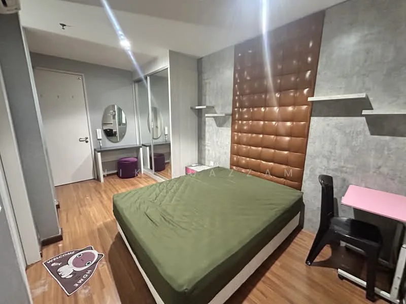 Bedroom features: queen-size bed with green bedding, brown upholstered accent wall, wooden flooring, built-in wardrobe with mirror, desk with pink tabletop, black chair, wall-mounted shelves, round mirror, decorative rug with cartoon character; style: modern.