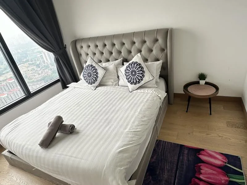 Bedroom features: king-size bed with upholstered headboard, white bedding with striped pattern, two decorative pillows with floral pattern, wooden bedside table, small potted plant on bedside table, area rug with tulip design, floor-to-ceiling window with city view, grey curtains, light wood flooring; style: modern; view: city.