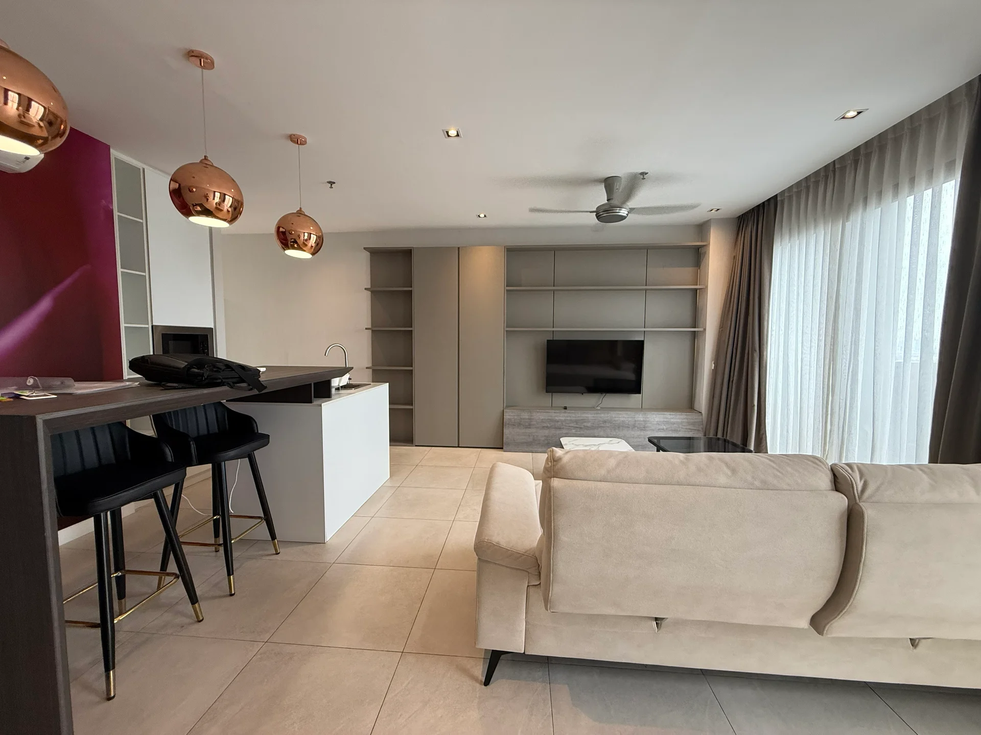 Living features: 3-seater fabric sofa in beige, coffee table, wall-mounted TV, ceiling fan, floor-to-ceiling windows with sheer curtains, light grey tiled flooring, decorative wall with pink accent, built-in shelving unit, open-plan layout with kitchen, recessed downlights; style: modern; view: city. Kitchen features: white kitchen island with sink, stainless-steel faucet, bar seating with black stools, built-in oven, microwave, dark cabinetry, light grey tiled flooring; style: modern.
