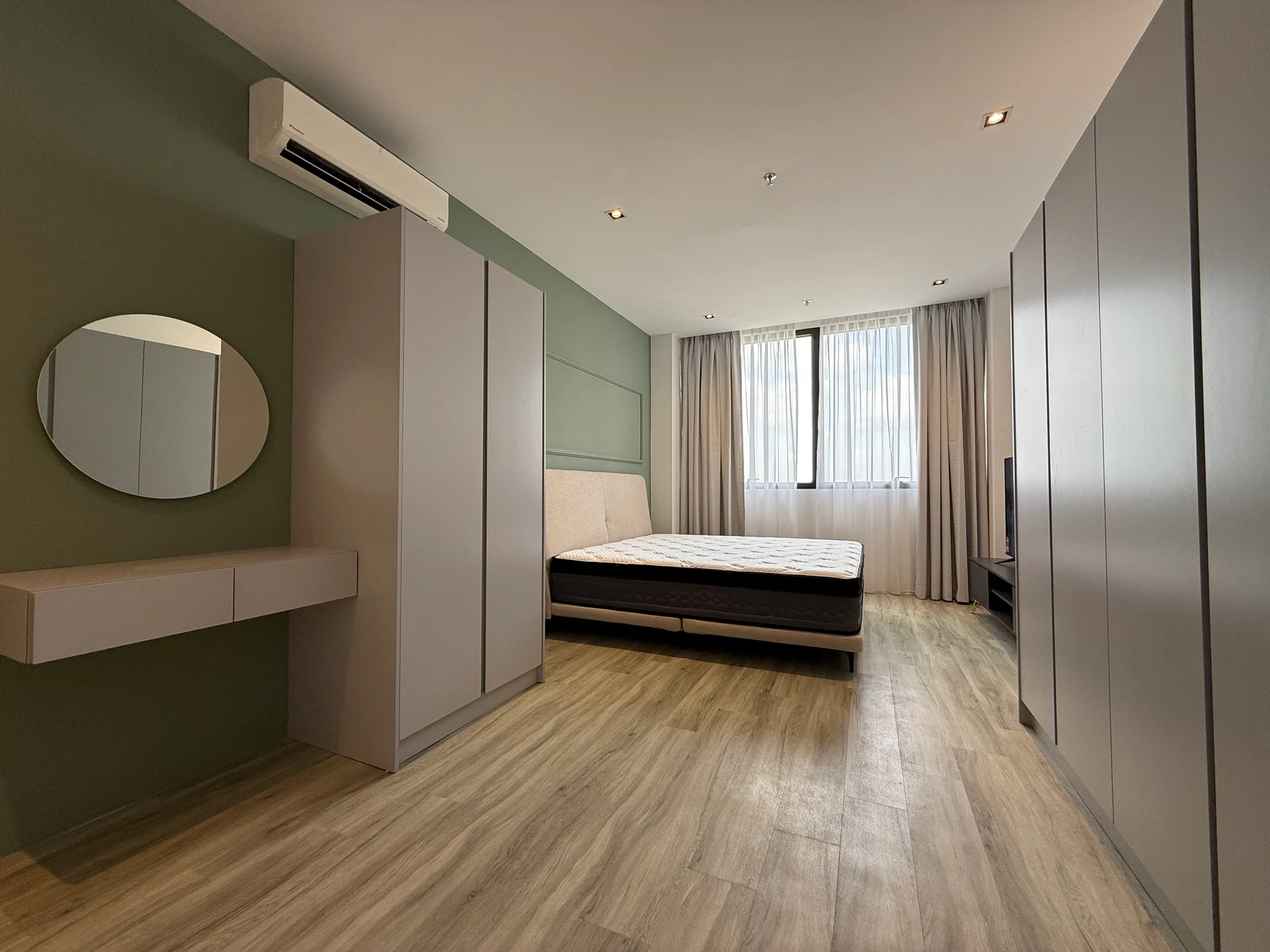 Bedroom features: wooden flooring, green accent wall, white walls, floor-to-ceiling window, sheer curtains, queen-size bed, upholstered headboard, built-in wardrobe with sliding doors, vanity table with mirror, wall-mounted TV, air conditioning unit, recessed downlights; style: modern.