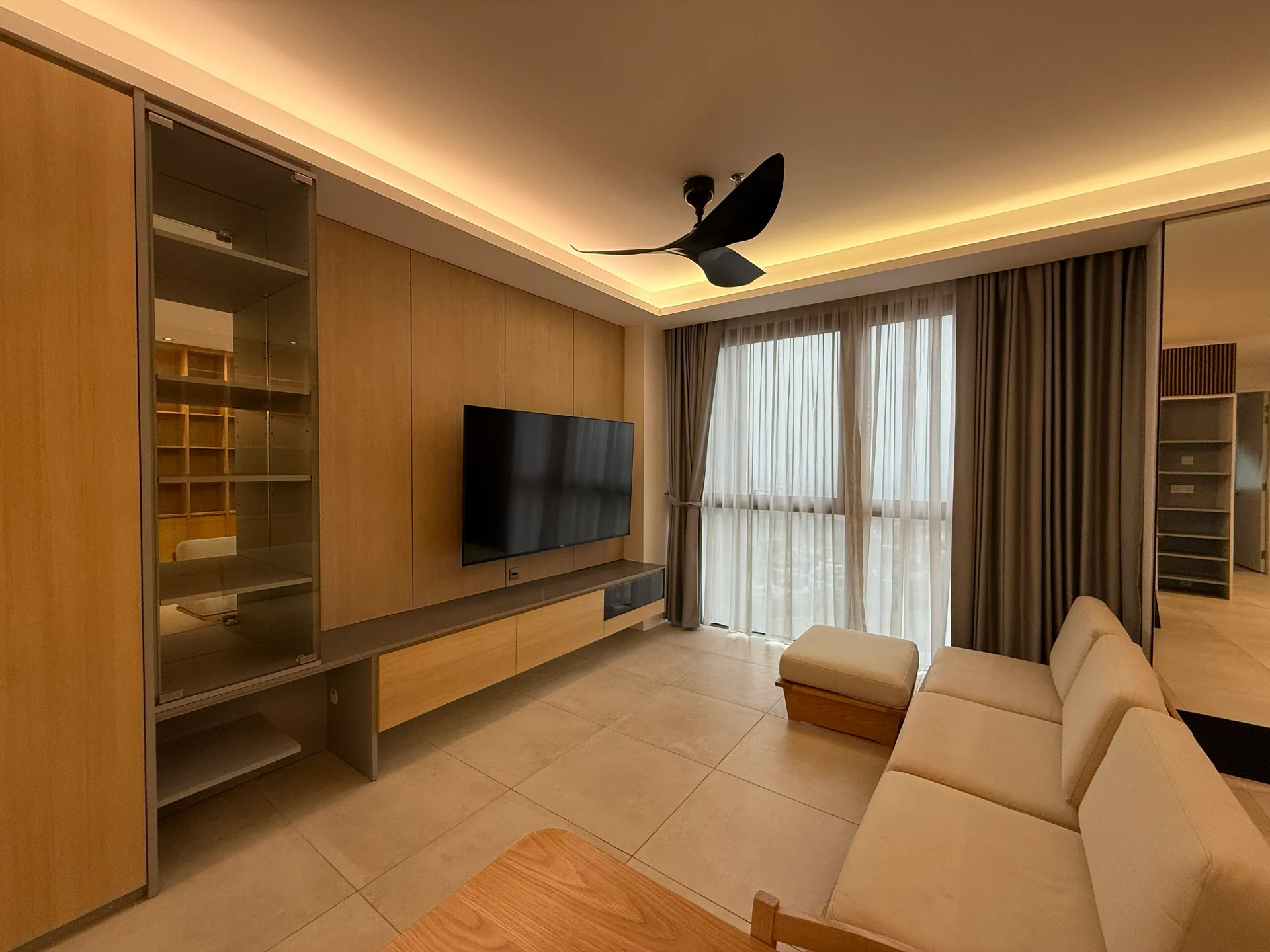Living features: light-coloured tiled flooring, built-in wooden cabinetry, wall-mounted flat-screen TV, 3-seater fabric sofa in light beige, ceiling fan with 3 blades, large floor-to-ceiling windows with sheer curtains, recessed LED lighting, wooden coffee table; style: modern.