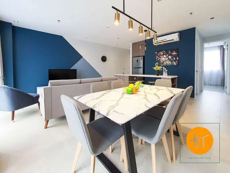 Dining features: marble dining table, 4 upholstered dining chairs in grey, pendant lighting above table, wall-mounted TV, blue accent wall, light grey walls, floor-to-ceiling windows, air conditioning unit, decorative wall art, open-plan layout to kitchen, neutral tile flooring; style: modern. Living features: 3-seater fabric sofa in light grey, coffee table, TV console, decorative plants, neutral tile flooring; style: modern. Kitchen features: stainless-steel fridge, built-in oven, freestanding range oven, kitchen cabinetry in light wood, countertop in light colour, open-plan layout to dining area; style: modern.